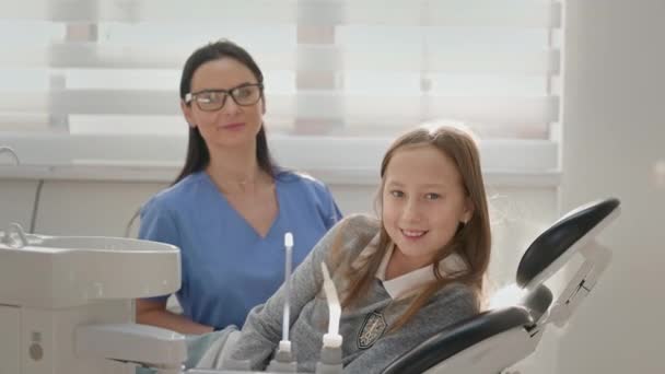 歯医者事務所の少女 長髪の可愛い女の子が歯科椅子に座っています 彼女の近くのメガネを持つ若い女性歯科医 小さな女の子の笑顔 — ストック動画