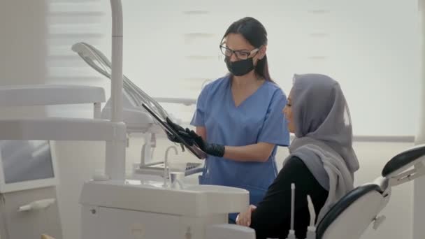 歯医者で働いている 若い患者の顔を閉じる 彼の頭の上にスカーフで若い女の子歯科医の椅子に座って マスクの若い女性医師は 患者のX線を示しています — ストック動画