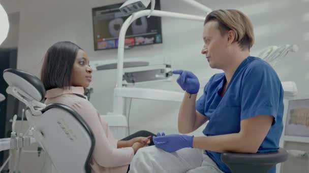牙科医生的接待处年轻男牙医与病人交谈 在椅子上坐着黑人少女 整整齐齐的医生办公室女孩笑了 — 图库视频影像