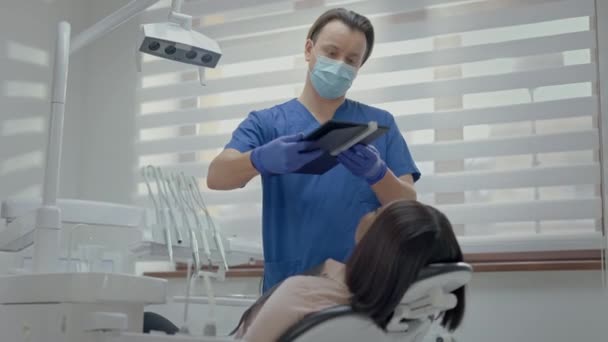 歯医者での受付 座長は黒人の少女 医師は タブレットを介して女の子の歯を見て 画面上の女の子の顔のイメージ 医療用手袋とマスクの医師 — ストック動画