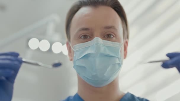 歯科室に立つ若い医師 ハンサムな男 顔を閉じてください 医者は医療マスクに直面する 手には医療器具を持っている 彼は治療を開始する患者に近づく — ストック動画