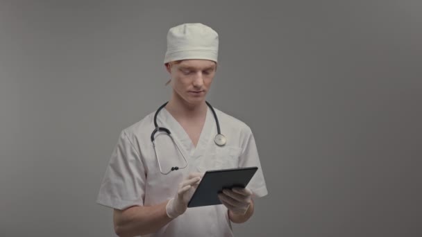 若い医師は 医療用のガウンと手袋彼は錠剤を保持します 首には聴診器 頭には白い帽子をかぶっている かわいい男は灰色の背景に立って — ストック動画