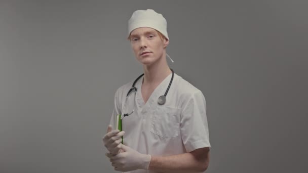 医療用ガウンと手袋の若い医師 首には聴診器 頭には白い帽子をかぶっている かわいい男は灰色の背景に立って — ストック動画