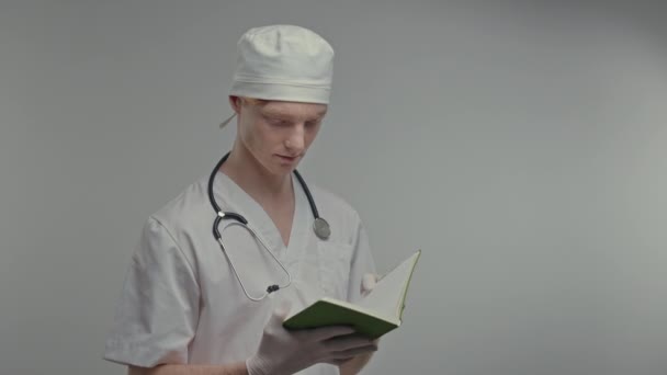 医療用ガウンと手袋の若い医師 首には聴診器 頭には白い帽子をかぶっている かわいい男は灰色の背景に立って — ストック動画