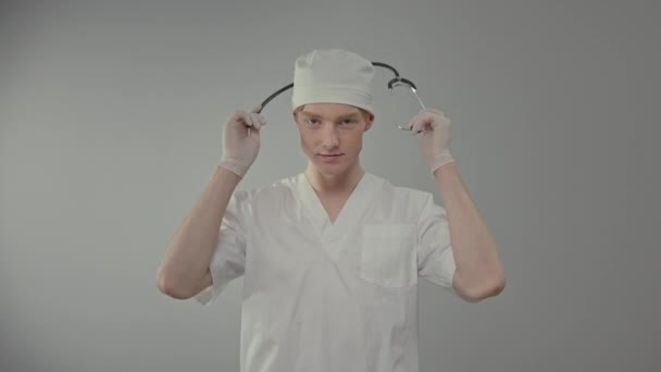 医療用ガウンと手袋の若い医師 かわいい男は灰色の背景に立って — ストック動画