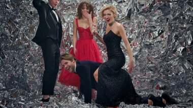 Four Young People At Party. Two Men In Suits And Two Girls In Evening Dresses. Brunette In Red Dress And Blonde In Black. Blonde Sitting On Back Of Guy With Glass Of Champagne. Another Couple Is