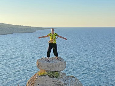 Siyah geniş pantolonlu ve sarı tişörtlü genç bir erkek saksafoncu denizin arka planında bir uçurumun tepesinde duruyor ve bir saksafon enstrümanı tutuyor. Aşırılık.