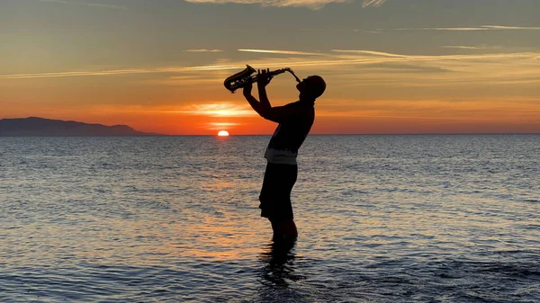 若い男性のサックス奏者は海の水の中で彼の足で立って 彼の手にサックスを保持し 日没を見ています 海の上に美しい夕日 ミュージシャン サックスを演奏 ダンス 楽しみを持って Silhouette — ストック写真