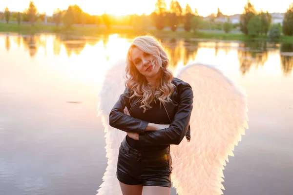 黒い革のジャケットを着たセクシーなブロンドの女性と白い天使の翼を持つショートパンツ 地獄や天国の悪魔や天使 湖の近くの夕日 — ストック写真