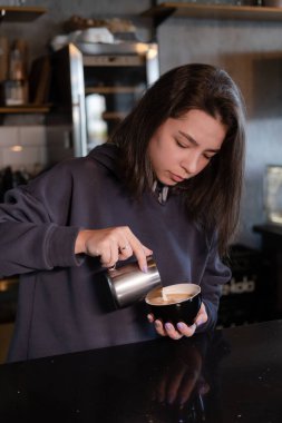 Şirin kız kahvecide latte yapıyor. Barista kahve yapıyor. İçecekte sütlü bir resim çizer - kapuçino.