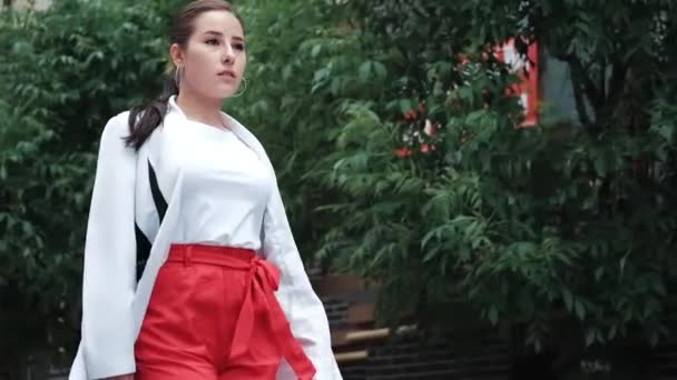 빨간 바지에 흰 블라우스와 재킷을 입은 자신있는 갈색 머리 여자가 거리를 걷고 있습니다. 느린 동작 — 비디오