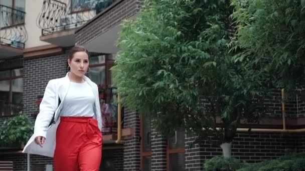 Pewna siebie brunetka w czerwonych spodniach i białej bluzce i kurtce spacerująca po ulicy. zwolniony ruch — Wideo stockowe