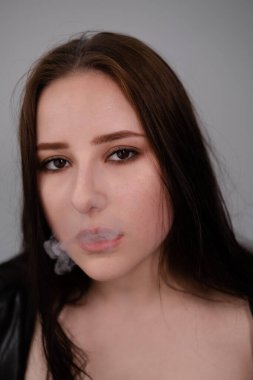 Siyah deri ceketli esmer kadın beyaz duman üflemiş. Gri arka planda sigara içen güzel bir kız. Kötü alışkanlık.