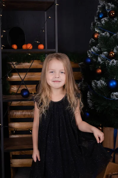 Charmig förskola blond flicka i en svart klänning står nära en julgran med blå dekorationer i mörkt rum — Stockfoto