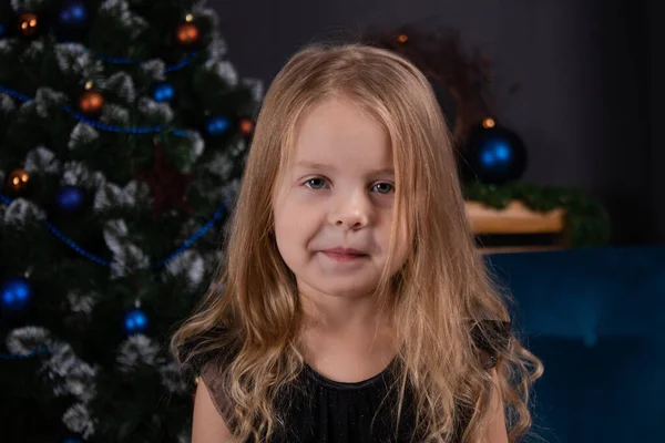 검은 드레스를 입고 어두운 방에 파란 장식 이 있는 크리스마스 트리 근처에 서 있는 매력적 인 미취학 전 금발 소녀 — 스톡 사진