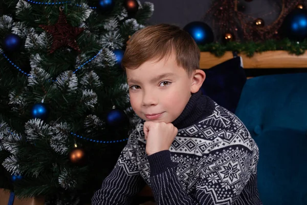 Okouzlující blondýnka dospívající chlapec v pletený tmavě modrý svetr v blízkosti zdobené vánoční stromeček. Veselé svátky. předvečer nových let — Stock fotografie