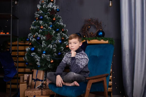 Portret chłopca w swetrze w ozdobionym pokoju z choinką i prezentami. ciemnoniebieskie kolory — Zdjęcie stockowe