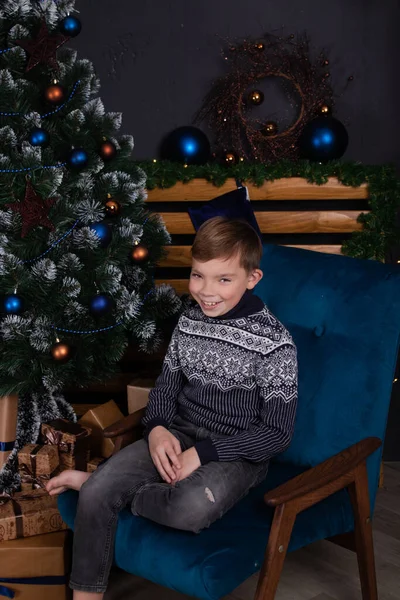 Encantador adolescente rubio en un jersey azul oscuro de punto cerca del árbol de navidad decorado. Felices fiestas. año nuevo víspera —  Fotos de Stock