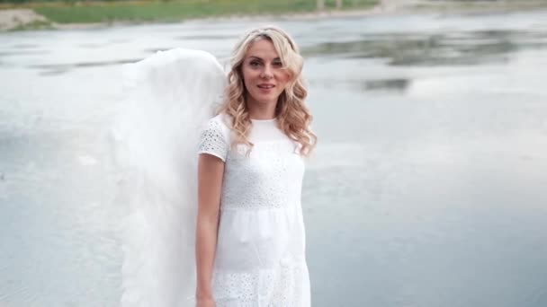 하얀 천사 날개를 가진 매력적 인 금발의 금발 이 물 옆에 서 있다. 천사 복장을 한 아름다운 여인. 여신. 느린 동작 — 비디오