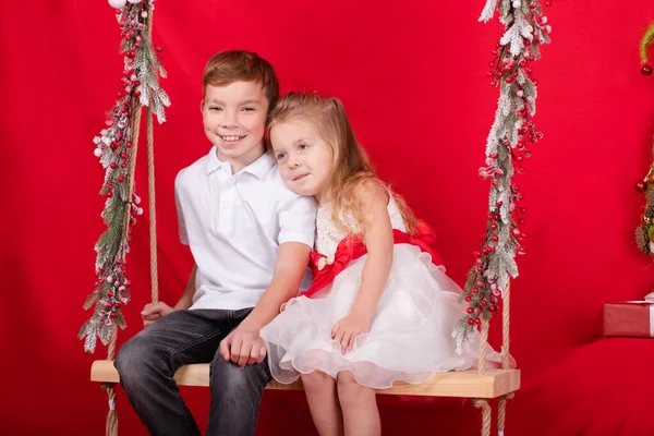Söt blond pojke och flicka sitter på en gunga dekorerad med trädgrenar och jul inredning. om röd bakgrund — Stockfoto