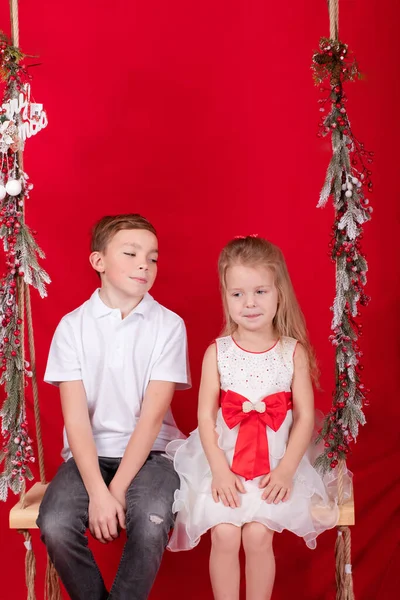 예쁜 금발의 소년 과 소녀가 나뭇가지로 장식된 그네에 앉아 크리스마스 장식을 하고 있습니다. 빨간 배경 — 스톡 사진