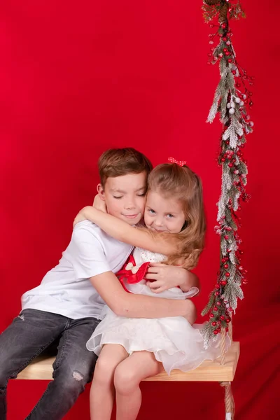 Familjetid. pojke och flicka - syster och bror - syskon inomhus nära en gunga dekorerad med juldekor och trädgrenar. röd bakgrund. nytt år tema — Stockfoto