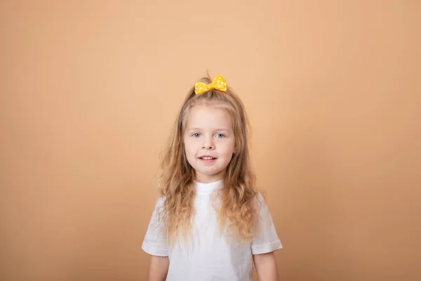 Portré egy édes szőke lányról fehér pólóban, sárga masnival a fején. barna alapon — Stock Fotó