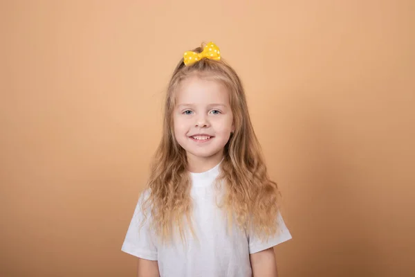 흰색 티셔츠를 입고 노란 활을 쓴 예쁜 금발의 여자 아이의 사진. ( 영어 ) 갈색 배경 — 스톡 사진
