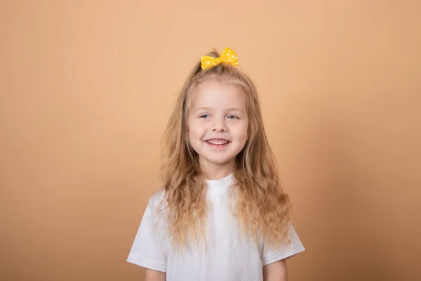 Portrait d'une douce petite fille blonde en t-shirt blanc et avec un nœud jaune sur la tête. sur fond brun — Photo