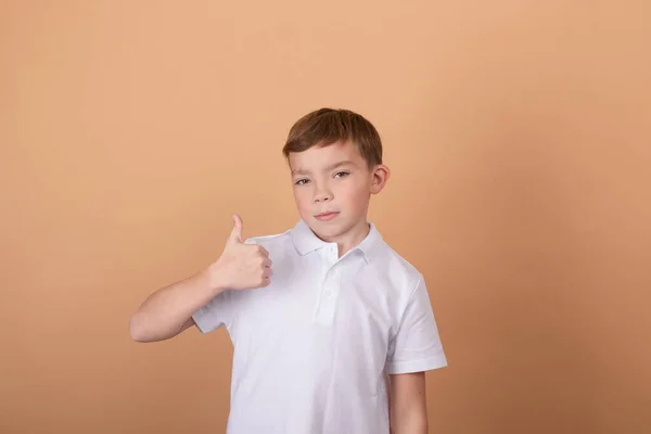 Jól mennek a dolgok. Csinos iskolás fiú fehér pólóban a kamerába néz, és mutogatja a hüvelykujját fényes barna háttérrel. Portré jóképű tinédzser srác 9-11 éves hasonló felirattal — Stock Fotó
