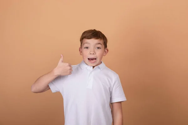 Portrét šťastného roztomilého dospívajícího chlapce v bílém tričku stojící a ukazující gesto palce nahoru, izolované na hnědém pozadí — Stock fotografie