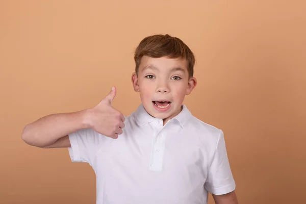 Portrét šťastného roztomilého dospívajícího chlapce v bílém tričku stojící a ukazující gesto palce nahoru, izolované na hnědém pozadí — Stock fotografie