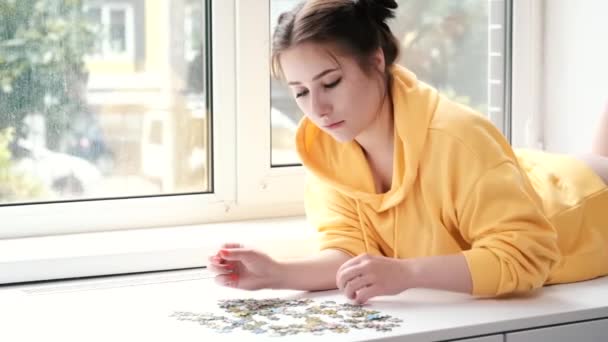 Bonita chica morena en sudadera con capucha amarilla la organización de piezas de un juego de rompecabezas en habitación blanca brillante. jugar tablero juego en casa. — Vídeos de Stock