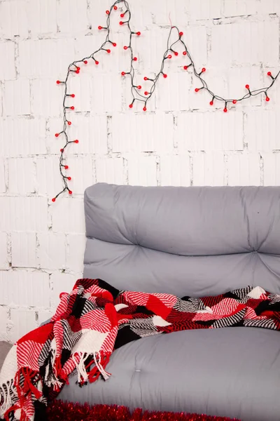 Gemütliche weihnachtliche Fotozone. graues Sofa mit rot karierter Decke. rote Girlande auf weißem Ziegelstein. rote Geschenkschachtel und Spielzeug Weihnachtsmann — Stockfoto