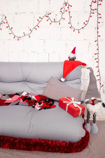 Útulná vánoční fotografická zóna. šedá pohovka s červenou kostkovanou přikrývkou. červené věnce na zdi z bílých cihel. červená dárková krabice a hračka Santa Claus — Stock fotografie