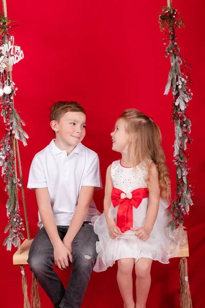 Хлопчик і дівчинка - сестра і брат - сидять на гойдалці, прикрашені різдвяним декором і гілками дерев. на червоному тлі. брати і сестри на Новий рік — стокове фото