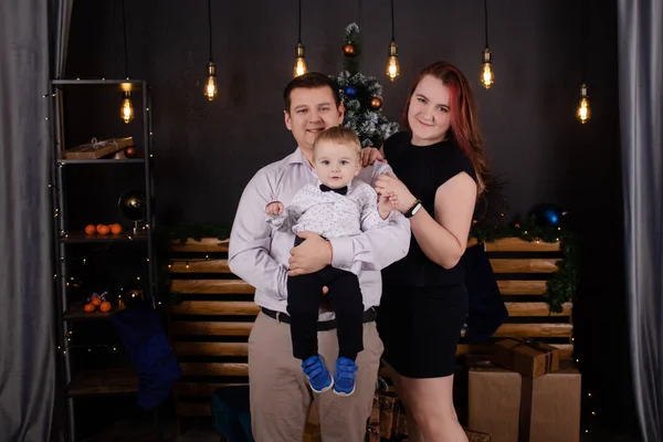 Father and mother holds their little blonde son and pose near the Christmas tree. Concept of family holiday. Young parents and baby boy have fun and hugs together in interior decorated for New year — Fotografia de Stock