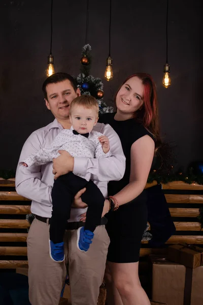 Father and mother holds their little blonde son and pose near the Christmas tree. Concept of family holiday. Young parents and baby boy have fun and hugs together in interior decorated for New year — Stockfoto