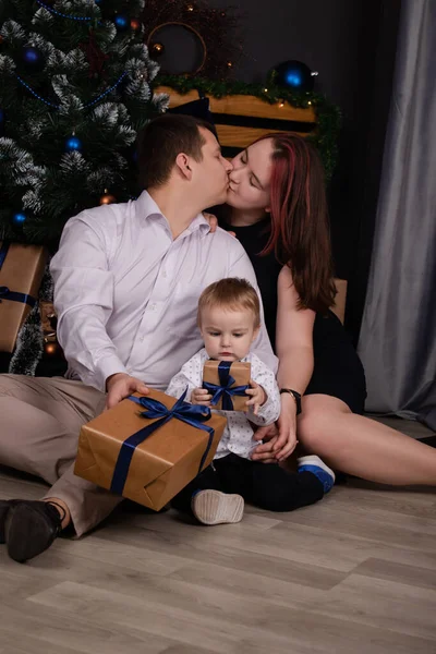 father and mother holds their little blonde son and pose near the Christmas tree. Concept of family holiday. Young parents and baby boy have fun and hugs together in interior decorated for New year