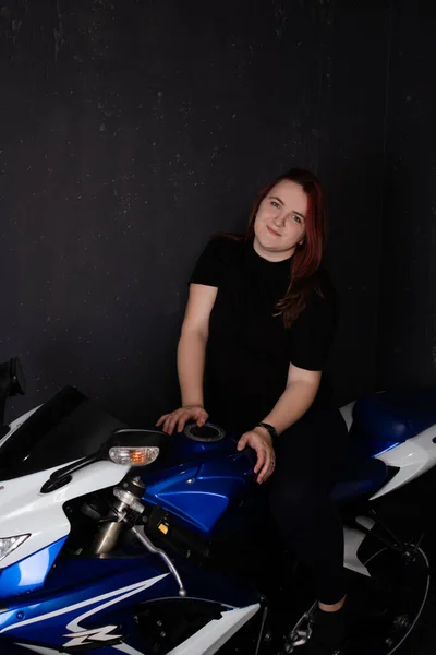 Redhead girl in black posing on blue motorcycle on black background. woman female moto-driver portrait — Stok fotoğraf
