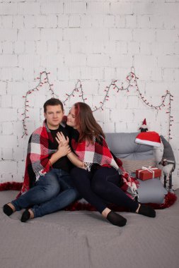 happy couple hugging on grey couch covered in red checkered plaid-blanket near red christmas decor. cozy young family