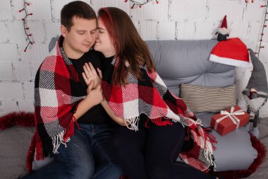 happy couple hugging on grey couch covered in red checkered plaid-blanket near red christmas decor. cozy young family