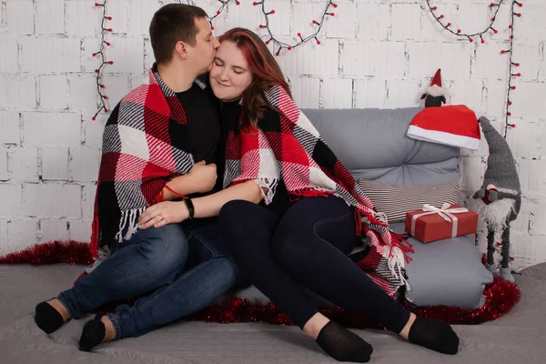 Feliz pareja abrazándose en el sofá gris cubierto de cuadros rojos manta a cuadros cerca de la decoración de Navidad roja. acogedora familia joven — Foto de Stock