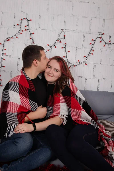 Feliz pareja abrazándose en el sofá gris cubierto de cuadros rojos manta a cuadros cerca de la decoración de Navidad roja. acogedora familia joven — Foto de Stock