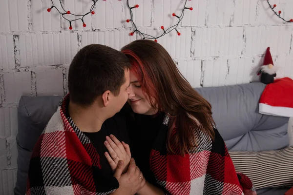 Щаслива пара обіймається на сірому дивані, вкритому червоною картатою плащеницею біля червоного різдвяного декору. затишна молода сім'я — стокове фото