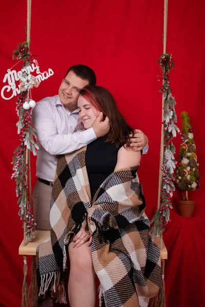 Pareja feliz abrazándose en columpio de madera decorado con ramas de árbol de Navidad sobre fondo rojo. acogedora familia joven — Foto de Stock