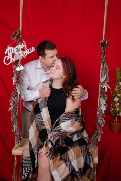 Happy couple hugging on wooden swing decorated with christmas tree branches on red background. cozy young family — Fotografia de Stock