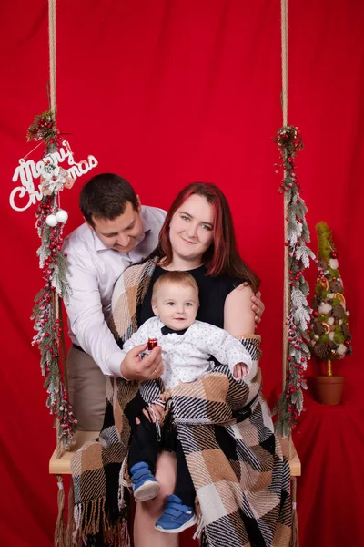 Happy family hugging on wooden swing decorated with christmas tree branches on red background. cozy young family. mother father and little baby son — Stock Fotó