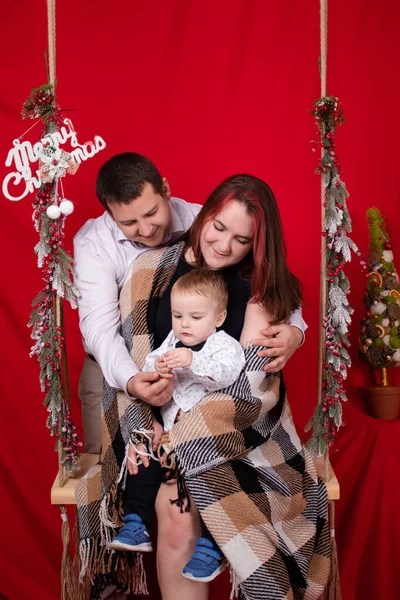 Happy family hugging on wooden swing decorated with christmas tree branches on red background. cozy young family. mother father and little baby son — Fotografia de Stock