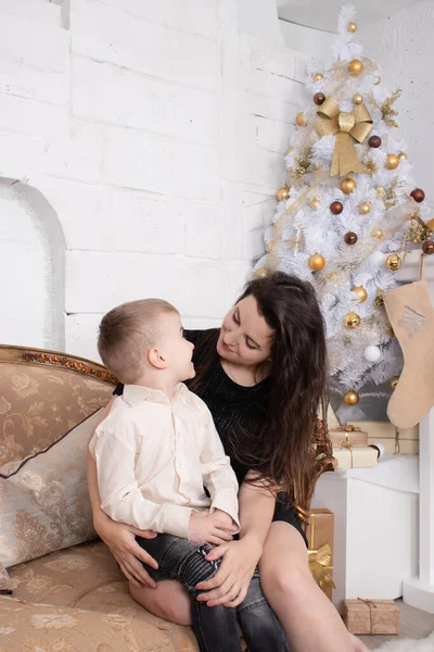Atmosphere of coming holidays. portrait of happy single mother with her little pleasant boy in white bright room near christmas tree in golden tones — Zdjęcie stockowe